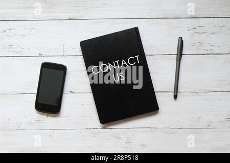 Top view mobile phone,pen and notebook written with CONTACT US on white wooden background. Stock Photo