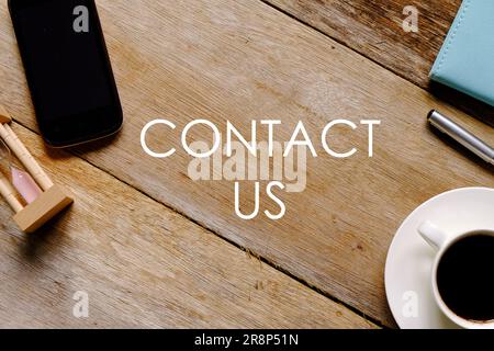 Top view of a cup of coffee,notebook,pen,mobile phone and hourglass on wooden background written with 'CONTACT US'. Stock Photo