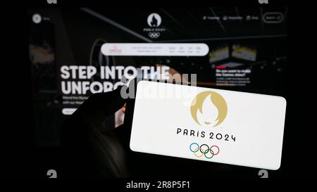 Person holding mobile phone with logo of the 2024 Summer Olympics in Paris on screen in front of web page. Focus on phone display. Stock Photo