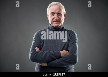 Smiling senior man with crossed arms wearing polo-neck and looking at camera in studio Stock Photo