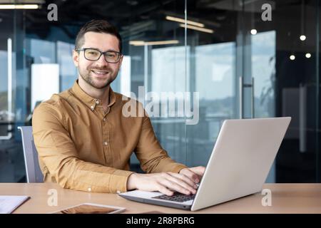 Portrait of a young male student confidently sitting at a desk in the office and smiling at the camera, learning remotely from a laptop. Stock Photo