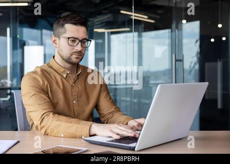 Portrait of a serious young male programmer, specialist developer who is concentrating on working on a laptop while sitting at a desk in an office center. Stock Photo