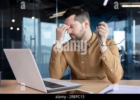 Young male businessman working overtime in the office on a laptop on a project, deadline. A tired person takes a break, holds glasses in his hands, massages his eyes with his hand, rests. Stock Photo
