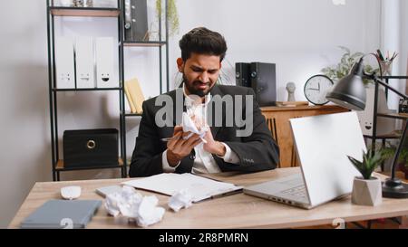 Angry furious indian man working at office throwing crumpled paper, having nervous breakdown at work, migaine, headache, stress management, mental distress problems, losing temper, reaction on failure Stock Photo