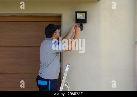 Image of an electrician on a scissor ladder using his tools to install a spotlight on a house. Illumination of the darkest parts of the house. Stock Photo