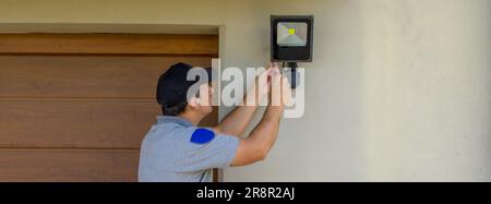 Image of an electrician on a scissor ladder using his tools to install a spotlight on a house. Illumination of the darkest parts of the house. Banner Stock Photo