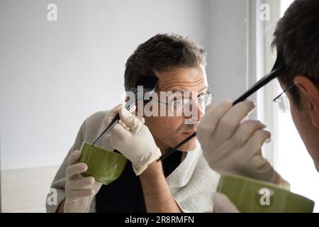 Middle-aged man with white hair is dyeing his hair in the mirror Stock Photo