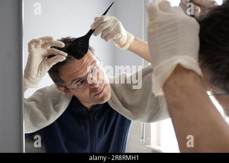 Middle-aged man with white hair is dyeing his hair in the mirror Stock Photo