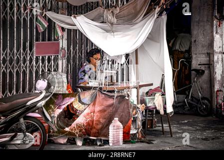 Bangkok, Thailand, March 20, 2023: Street garment mender working in front of a folding steel door and under a worn-out fabric sun shade. Stock Photo