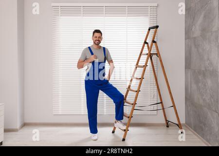 Worker in uniform and stepladder near horizontal window blinds indoors Stock Photo