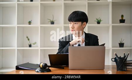 A professional and successful Asian male lawyer or legal counselor in a formal suit sits at his desk, looking out the window, and thinking or planning Stock Photo