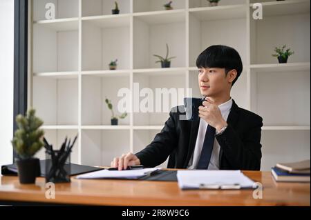 A handsome and successful millennial Asian male lawyer or legal consultant in a formal suit is sipping his morning coffee at his desk. Stock Photo