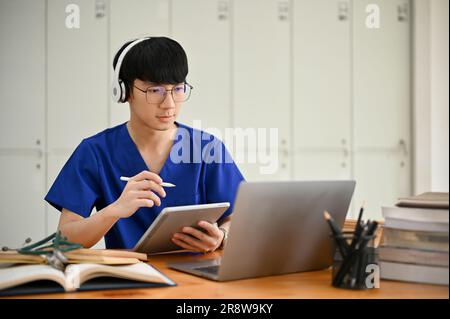 A smart and handsome young Asian male medical student in scrubs wearing headphones focuses on studying an online class through his laptop and taking n Stock Photo