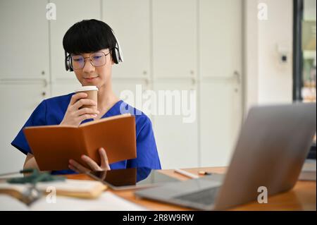 A smart and focused young Asian male medical student or doctor in scrubs wearing headphones, sipping coffee, and reading a book at his desk. Stock Photo