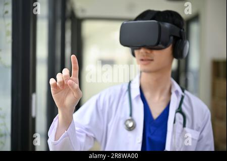 A smart and professional millennial Asian male doctor in a uniform wearing virtual reality goggles in the hospital corridor. Health care and technolog Stock Photo