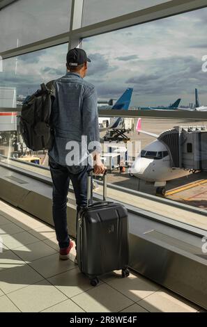 Man With Backpack, Suitcase standing in airport and looking at aircraft flight through window at Terminal. Man On His Way To Flight Boarding Gate. Vac Stock Photo