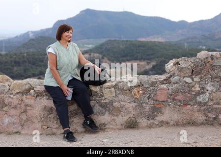 Active middle aged woman with backpack relaxes sits with water bottle on hill, looks at beautiful view after hiking up. Healthy lifestyle in retirement. Adventure seeks woman treks enjoys freedom. Stock Photo