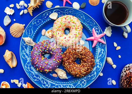 Assorted donuts in a plate on a blue background. Around them are symbols of the sea - shells, pebbles and stones. The concept of pleasure and relaxati Stock Photo