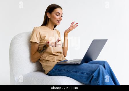 Cheery Caucasian woman in headphones having online video call on laptop, sitting on chair against white wall, full length. Millennial lady participati Stock Photo
