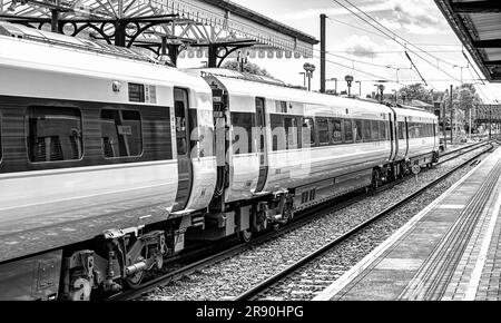 A line of railway carriages stand near a 19th Century historic canopy. An iron bridge is in the distance and a sky with cloud is above. Stock Photo