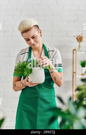 plant care, indoor gardening, smiling tattooed woman in green apron touching natural potted plant while standing in modern living room, sustainable ho Stock Photo