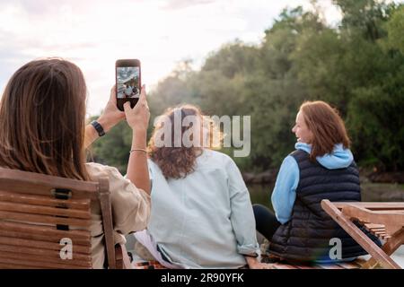 Women friends relaxing together outdoors in nature and taking photos using smart phone. Stock Photo
