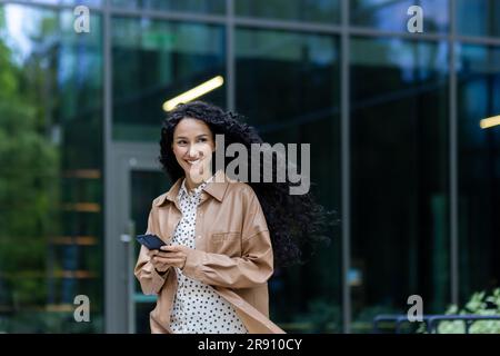 Young beautiful hispanic woman walking in the city, business woman holding phone in hands using smartphone app, woman smiling contentedly and happy outside office building with curly hair Stock Photo