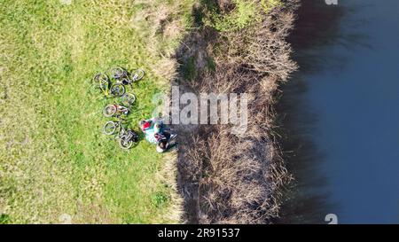 Family on bikes cycling outdoors, active parents and kids on bicycles, aerial view of family with children relaxing near beautiful river from above Stock Photo