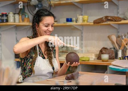 Young woman working raw ceramic and clay to make future ceramic product. Pottery workshop with natural light scraping, smoothing, shaping and sculptin Stock Photo