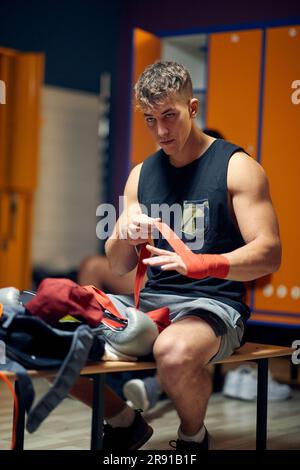 Young man in dressing room, sitting on bench, tying red tape around his hand, getting ready for workout. Boxing, martial arts, sports concept. Stock Photo