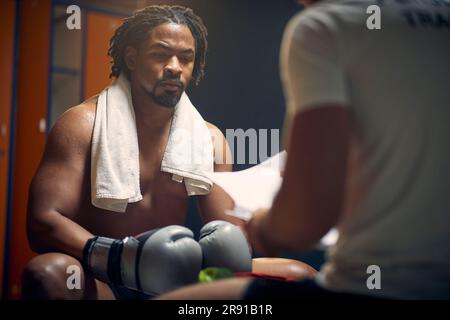Strong muscular boxer full of sweat after boxing workout resting, sitting in dressing room with his coach. Consulting with personal trainer. Stock Photo