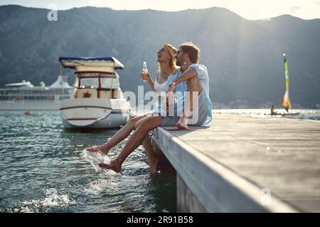 Couple having a drink and laughing by seaside sitting on wooden jetty by water. Holiday, lifestyle, togetherness concept. Stock Photo