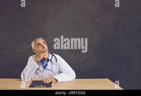 Portrait of dreamy or thinking senior male doctor with pensive expression on gray banner background. Stock Photo
