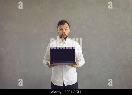 A young man stands with his mouth wide open, surprised, holding a laptop in his hands with a black screen. Stock Photo