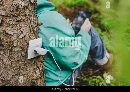 Man using tablet computer charging with outlet on tree trunk Stock Photo