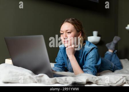Businesswoman in denim shirt working with laptop lying on bed Stock Photo