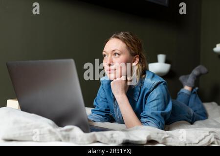 Thoughtful freelancer in denim shirt lying on bed with laptop Stock Photo