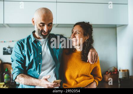 Man using smart phone by smiling woman in kitchen at home Stock Photo