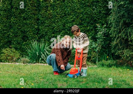 Mother teaching son to mow grass using toy lawnmower in garden Stock Photo