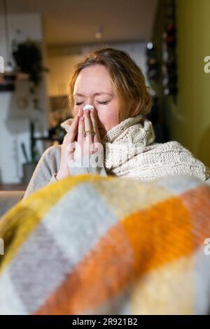 Sick young woman wrapped in blanket sneezing at home Stock Photo