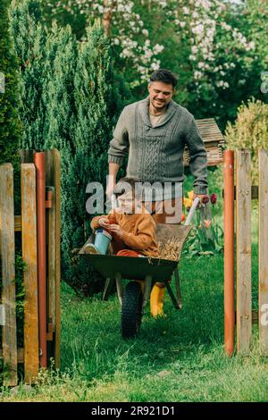 Father pushing son sitting in wheelbarrow at back yard Stock Photo
