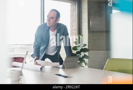 Thoughtful businessman leaning on table with paper and tablet PC in office Stock Photo