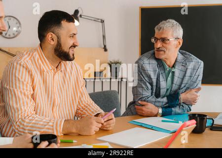 Male colleagues laugh and talk sitting at studio table Stock Photo