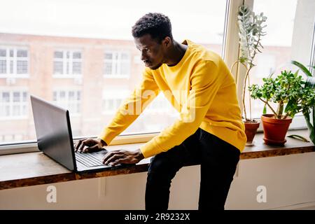 Focused man using laptop sitting on window sill at home Stock Photo