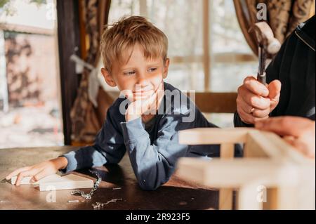 Son leaning on elbow looking at father making birdhouse at home Stock Photo