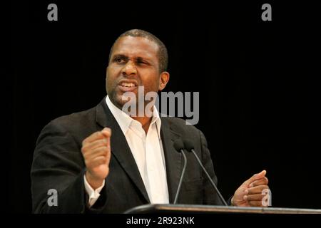 A young Caucasian man stands confidently in front of a microphone on a stage, addressing a crowd of listeners at a formal event Stock Photo