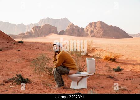 Contemplative man sitting on toilet seat and defecating in desert Stock Photo