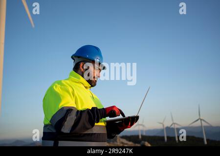 Engineer wearing hardhat using laptop near wind turbines Stock Photo