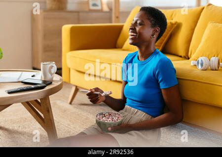 Cheerful young woman eating cereals and watching TV at home Stock Photo