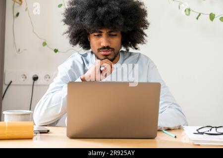 Thoughtful businessman with hand on chin working on laptop at home Stock Photo